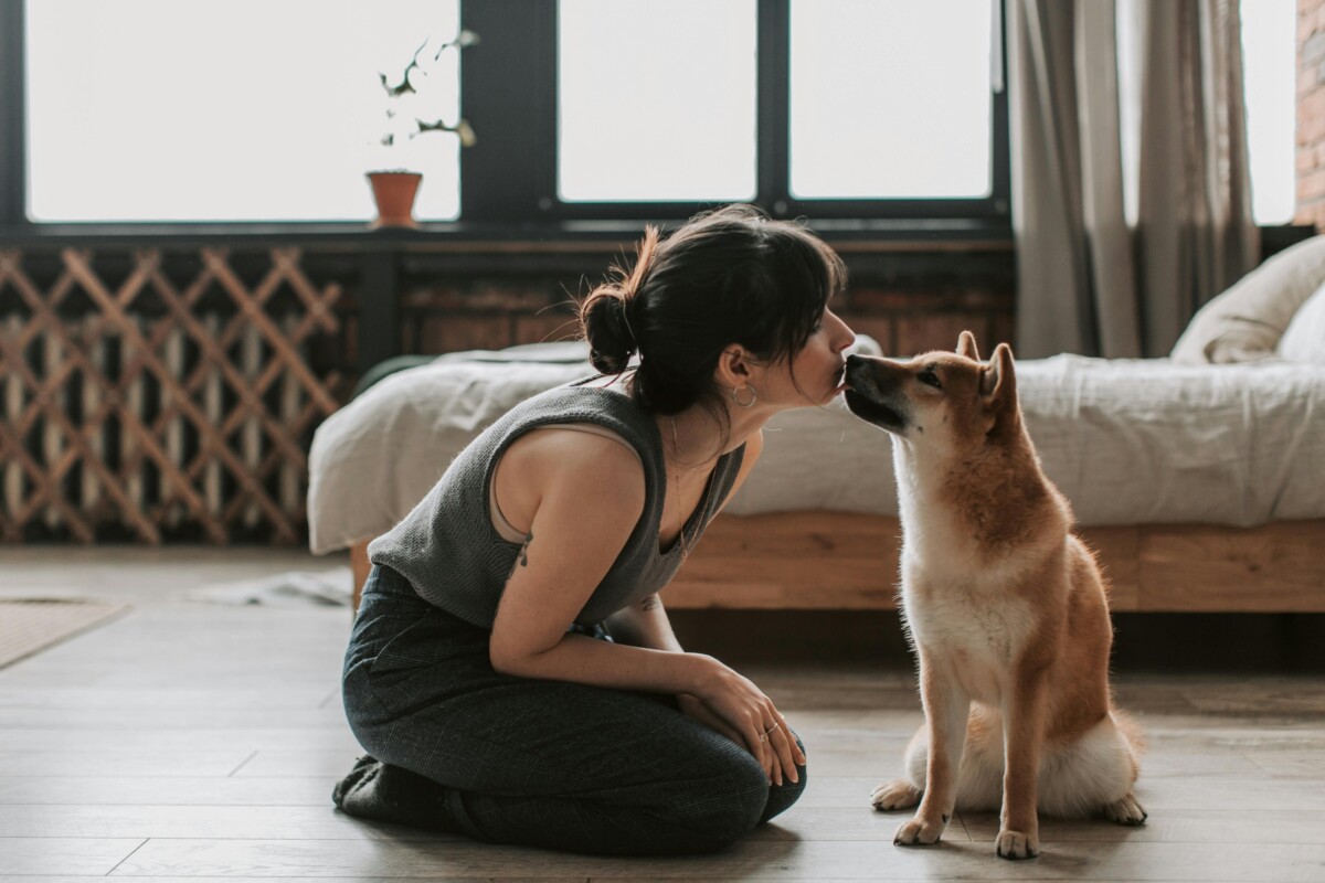 A woman kneeling on the floor and sharing a gentle nose-to-nose moment with her dog in a cozy bedroom setting.